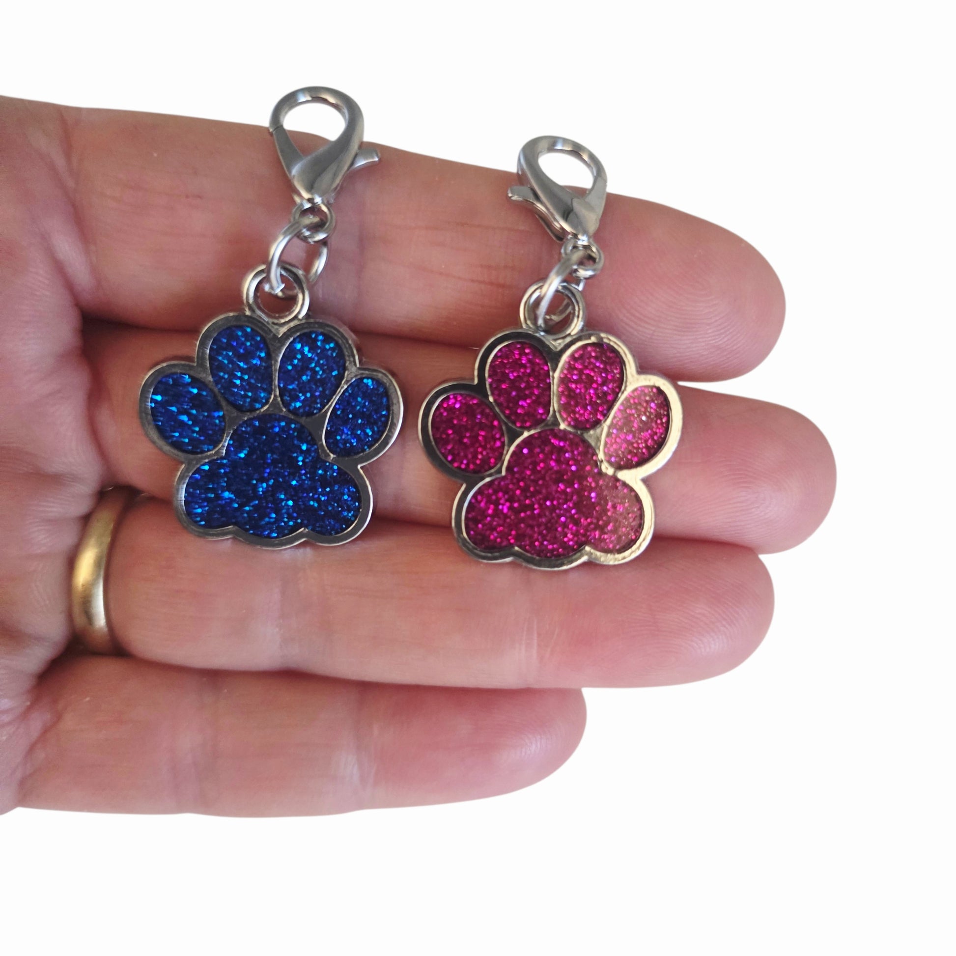 Two enamel paw charms, one blue and one pink, held between fingers on a white background.