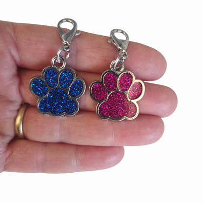 Two enamel paw charms, one blue and one pink, held between fingers on a white background.