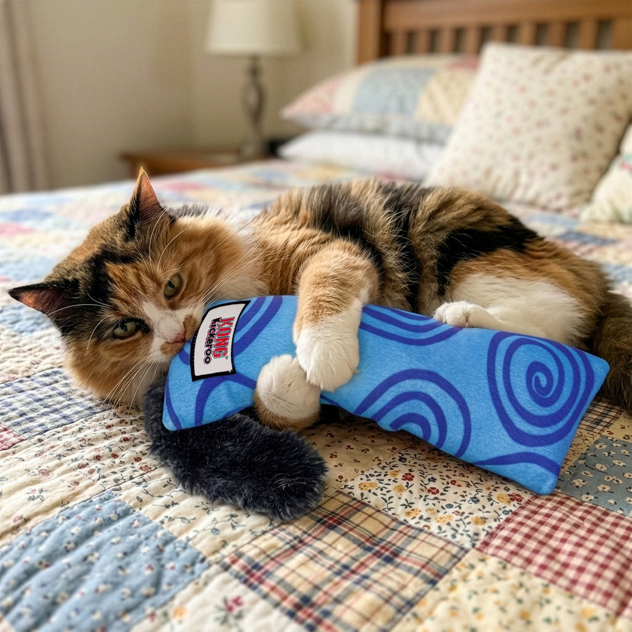 Cat playing with a blue KONG Kickeroo Swirl cat toy on a patchwork quilted bed.