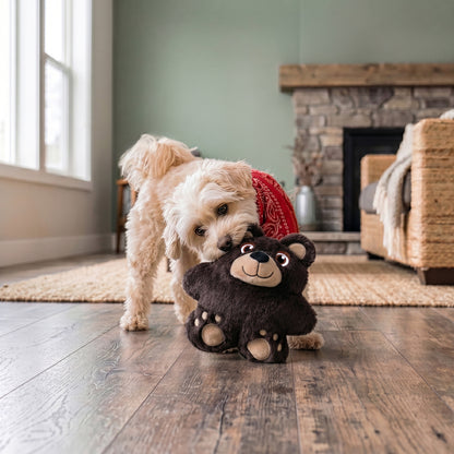 Dog playing with a plush KONG Snuzzles bear toy on a wooden floor in a cozy living room.