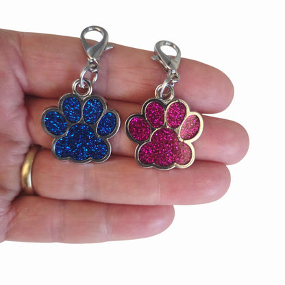 Two glittery paw-shaped charms, one blue and one pink, held between fingers on a white background.