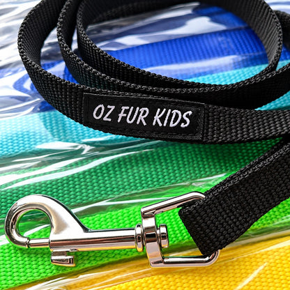A close-up of a black woven dog leash with a silver metal clasp, laid over a colorful packet of leads. The leash has a black fabric label with white text reading 'OZ FUR KIDS'.